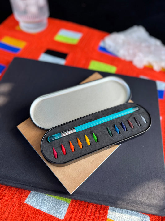 Set of color pencil and color pencil nibs in a black case on a colorful surface with a glass, crystal, and books in the background. 