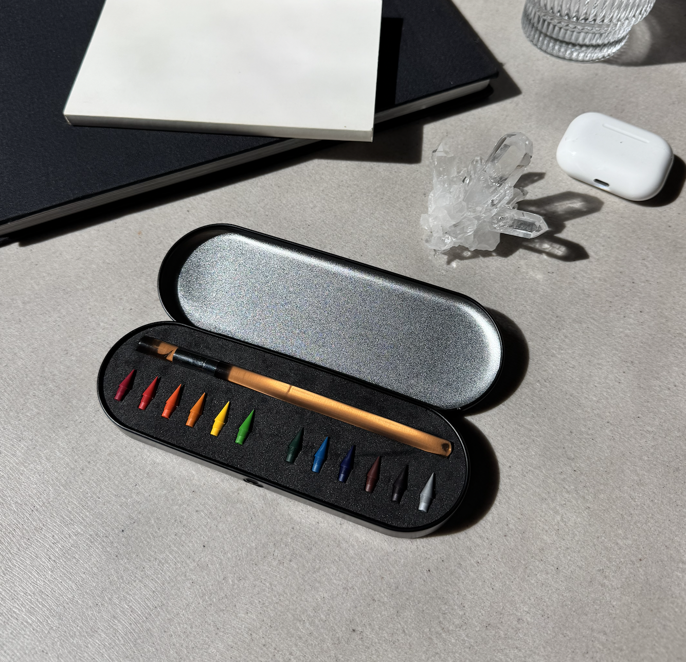 Set of color pencil and color pencil nibs in a black case on a light gray surface with a glass and books in the background. 