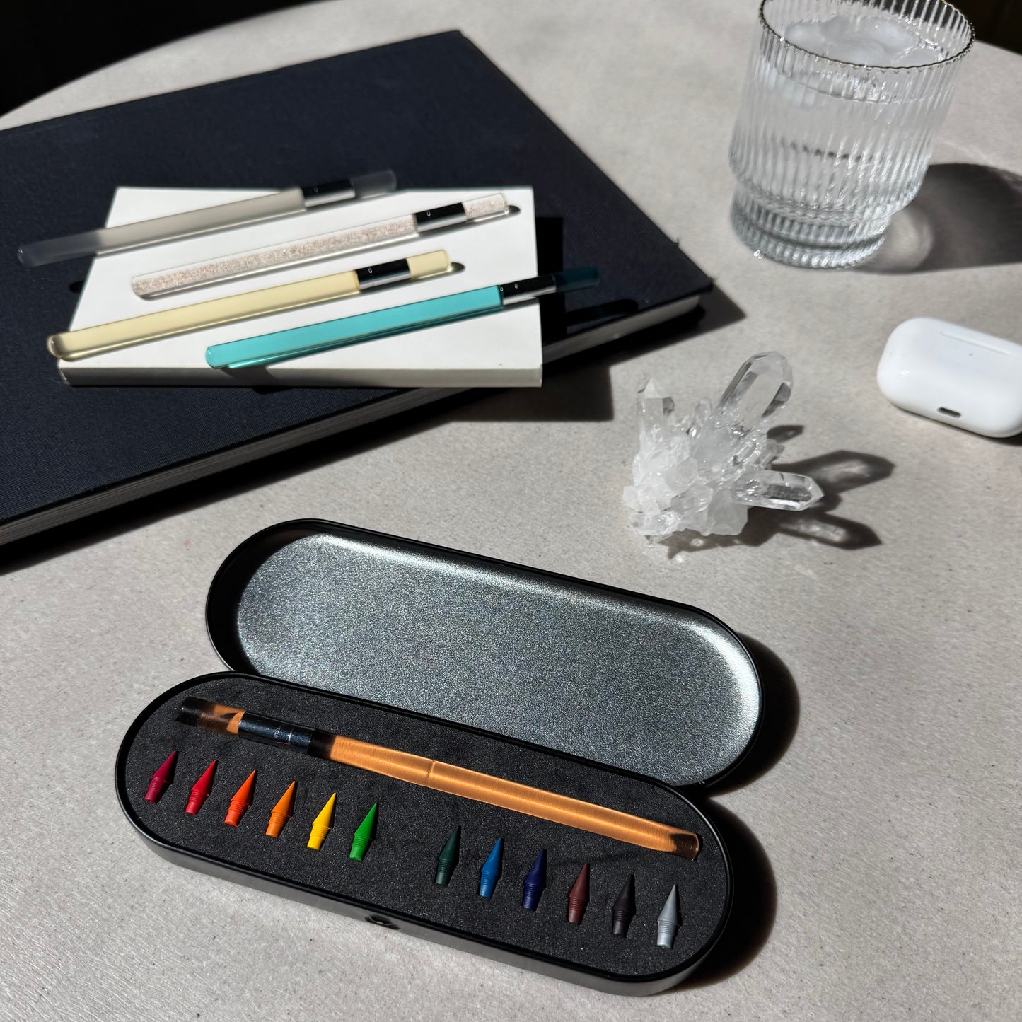 Set of color pencil and color pencil nibs in a black case on a light gray surface with a glass and books in the background. 