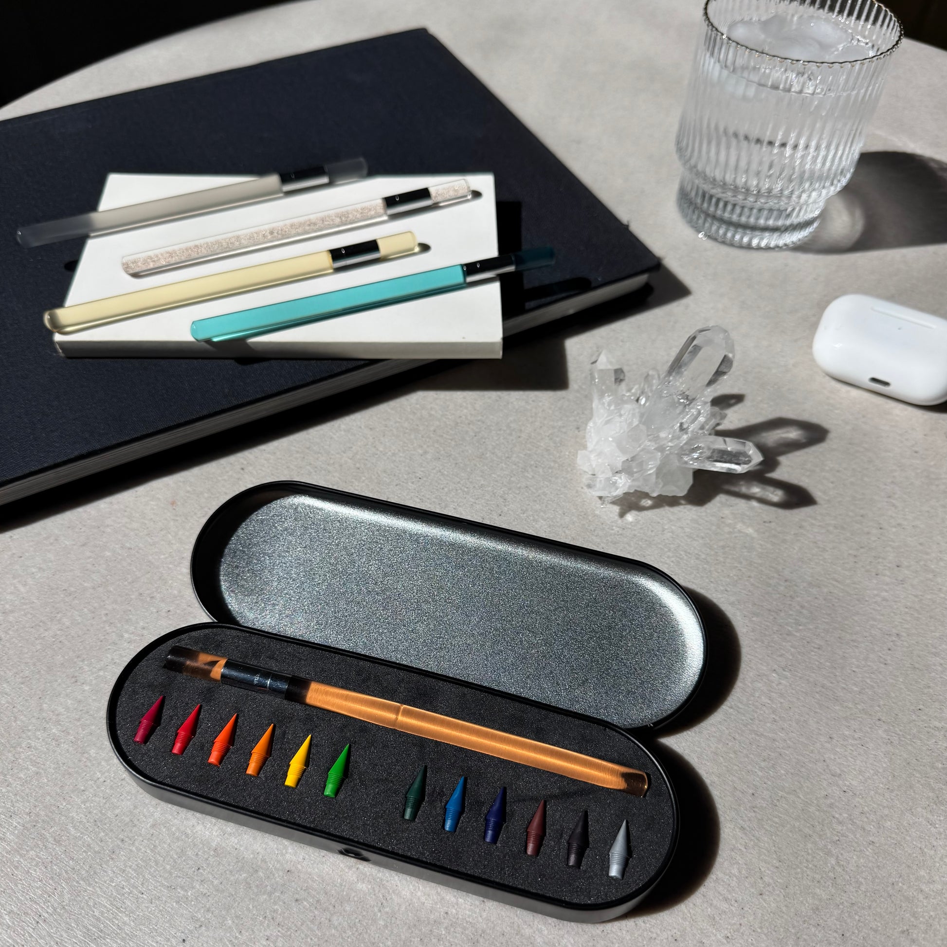 Set of color pencil and color pencil nibs in a black case on a light gray surface with a glass and books in the background. 